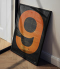Tilted framed McLaren poster with orange ring design leaning against a wall in a room