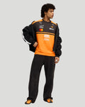 Full body of a male model wearing the McLaren Papaya 2026 Team T-Shirt with orange and black design, jeans and sneakers, jacket draped over shoulders.
