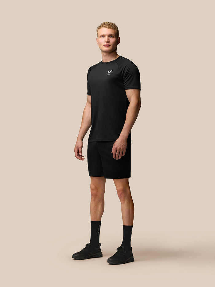 Full body shot of a man in black Adapt Training T Shirt with raglan sleeves and Castore logo on left chest against a beige background
