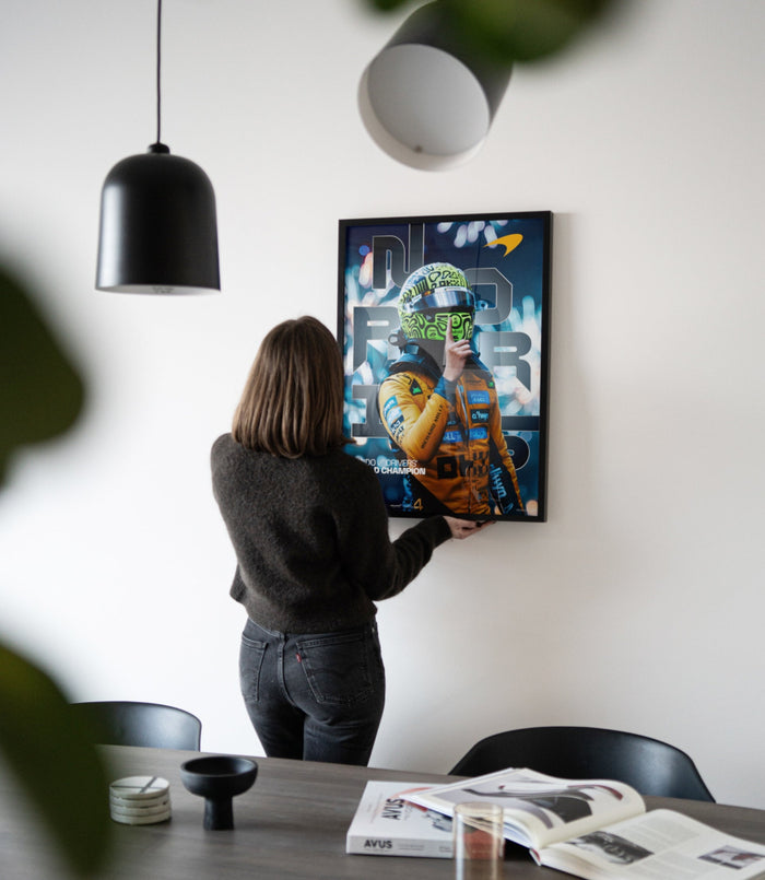 Woman in a dark sweater adjusts the framed Lando Norris 2025 World Drivers Championship poster on a textured wall above a desk.