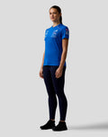 Side profile of a woman in a royal blue BWT Alpine Formula One Team Set Up T Shirt with front logos and pink piping along the shoulder