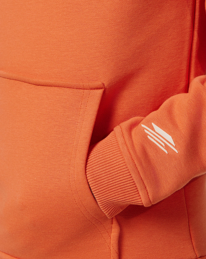 Close up of orange rust hoodie fabric featuring a stitched front pocket seam and a white diagonal logo on the sleeve.