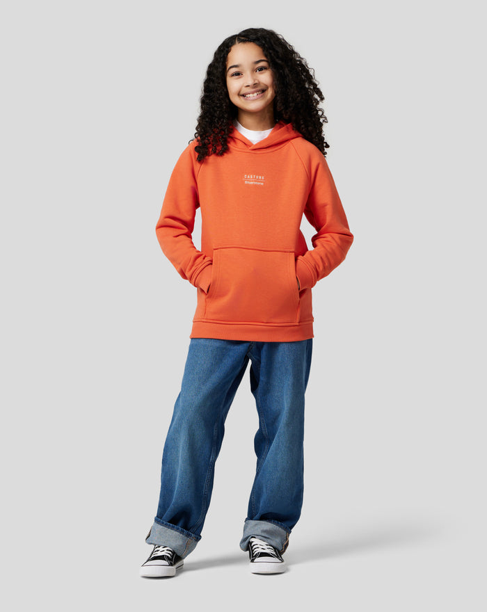 Child model wearing orange rust Junior Silverstone x Castore Hoodie with chest logo and front kangaroo pocket