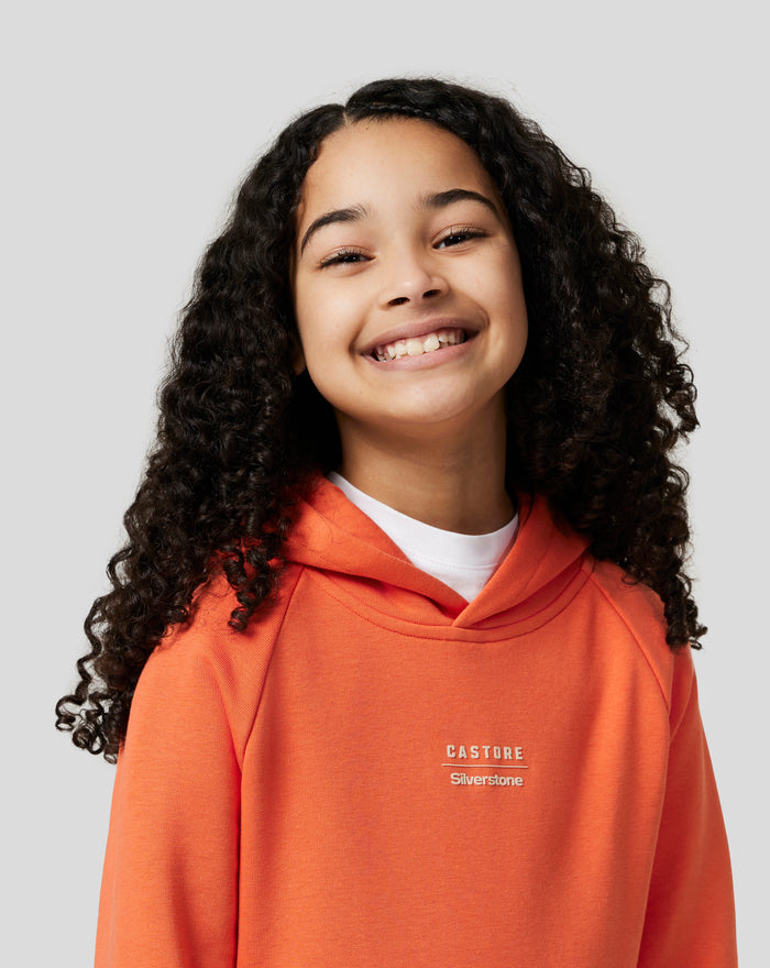Smiling young person wearing an orange rust junior hoodie with Castore Silverstone branding on chest.
