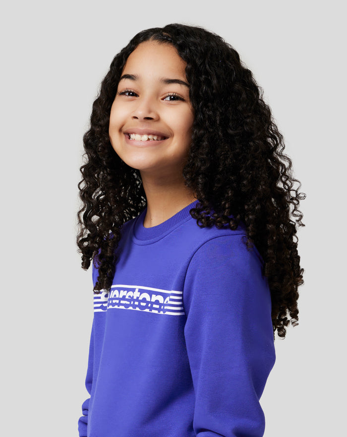 Close up of a smiling junior girl wearing a royal blue Junior Silverstone Core Sweatshirt against a light gray background