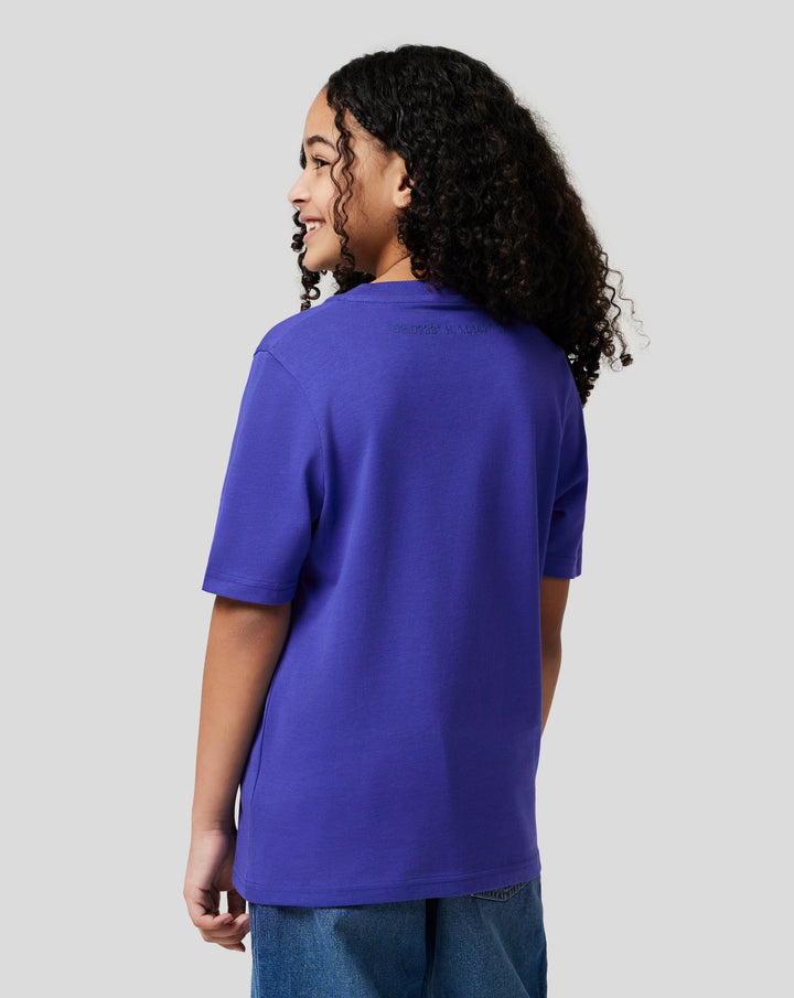 Side view of a young model wearing a royal blue Junior Silverstone Core Graphic T Shirt against a light gray background.