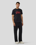 Full body shot of a man in a black Silverstone Legend T-Shirt with red graffiti logo, dark jeans and beige sneakers.