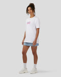 Female model wearing bright white festival graphic T shirt with pink text, paired with a light denim mini skirt and white sneakers.