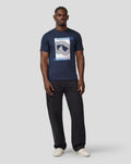 Model wearing the navy Core Track Graphic T Shirt with a bold central graphic and blue striped border, paired with dark jeans.
