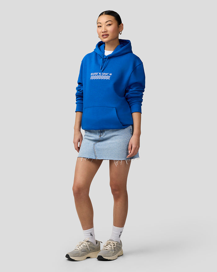 Three-quarter view of a woman in a blue Silverstone Core Hoodie with a white coordinates graphic on the chest