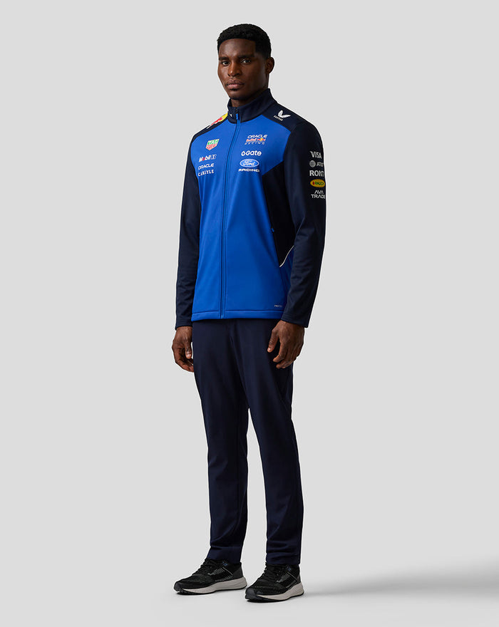 Full body view of male model wearing blue Oracle Red Bull Racing Teamline Soft Shell Jacket with sponsor logos on chest and sleeves.