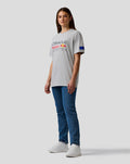 Full-body side view of a woman wearing a grey marl Oracle Red Bull Racing T-shirt with jeans and white sneakers.