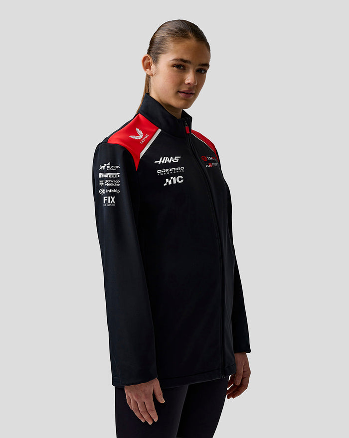Female model wearing Haas Team unisex soft shell jacket in black with red shoulder panels and logos on chest and sleeve.