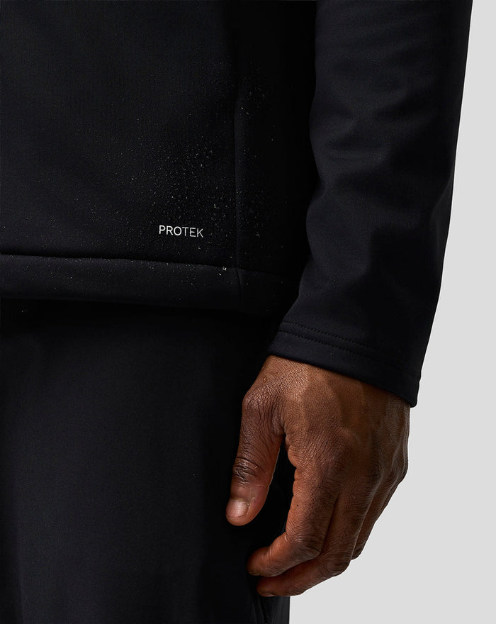 Close up of Haas Team Soft Shell Jacket showing PROTEK wordmark at the front lower left hem on black fabric.