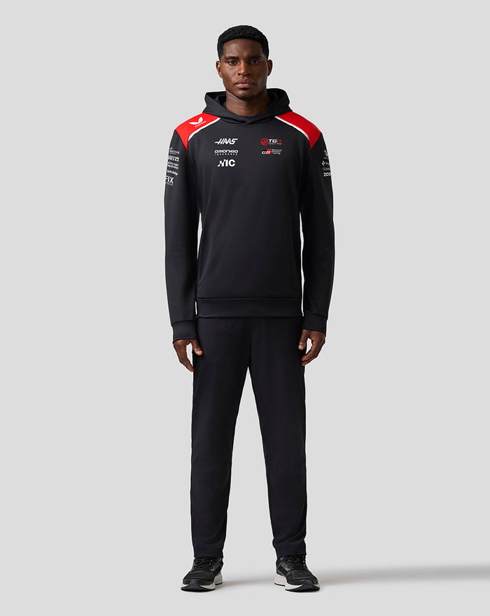 Full front view of a man wearing a black Haas Team pullover hoodie with red shoulder panels and sponsor logos on the chest.