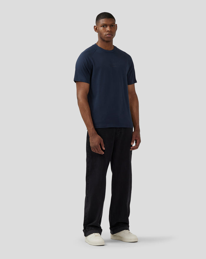 Full body shot of model in navy Silverstone x Castore T-shirt with embossed chest logo, dark jeans and white sneakers.