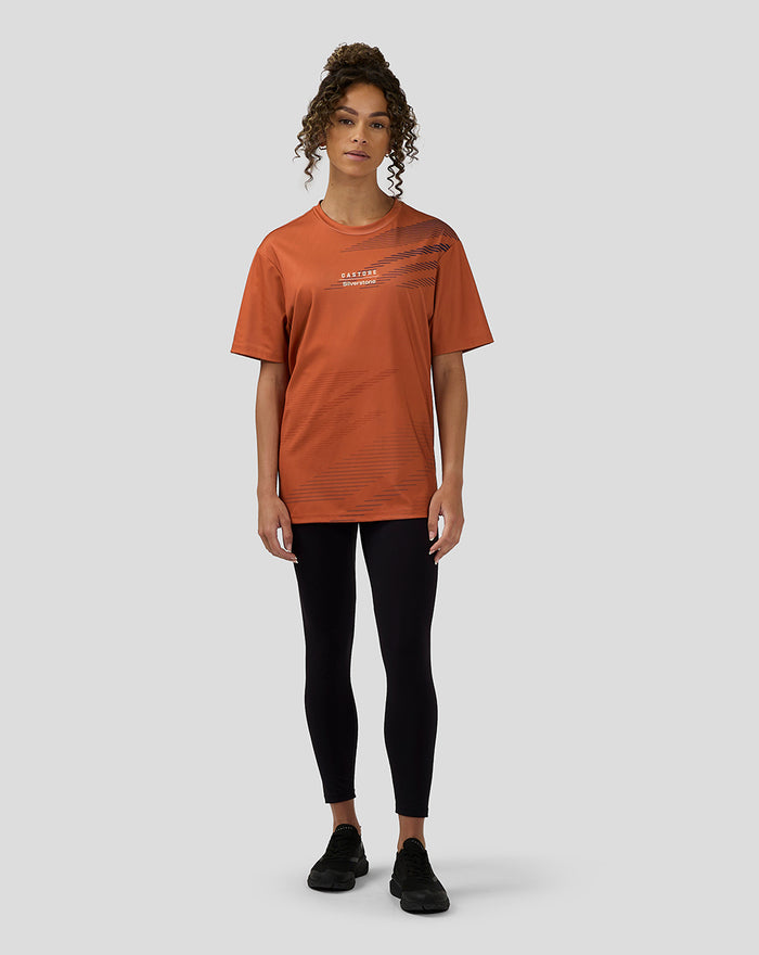 Full body shot of a woman in an orange rust Silverstone sublimated short sleeve T shirt with diagonal navy print black leggings and sneakers