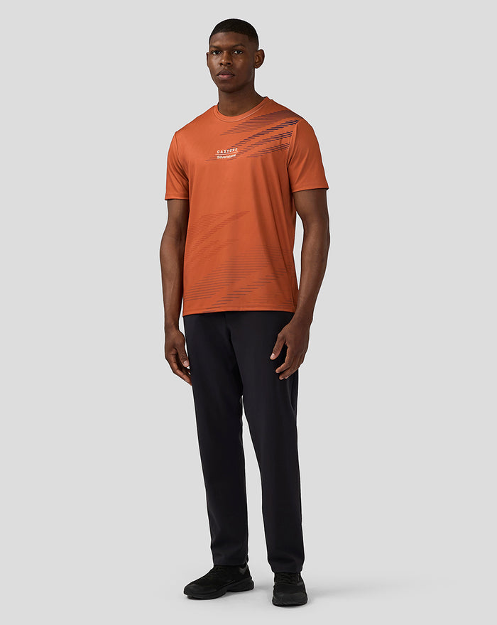 Full body shot of a male model in an orange rust Silverstone sublimated short sleeve T shirt with diagonal navy print and black pants