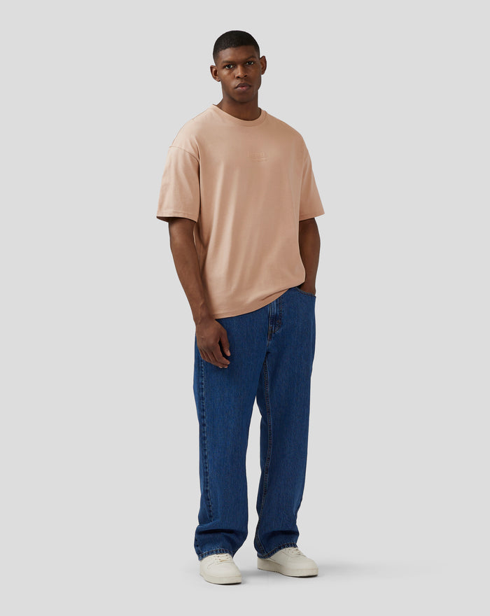 Male model wearing beige oversized short sleeve t shirt with embossed chest logo, blue jeans and white sneakers
