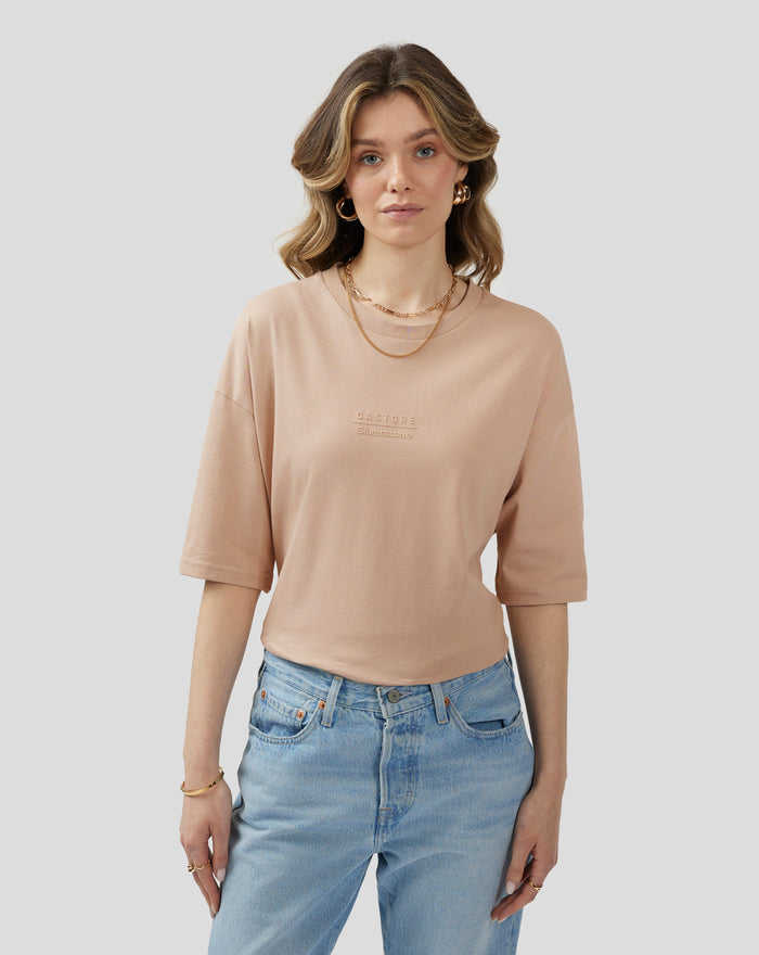 Female model wearing beige doeskin oversized short sleeve t shirt with embossed chest logo and back neck logo