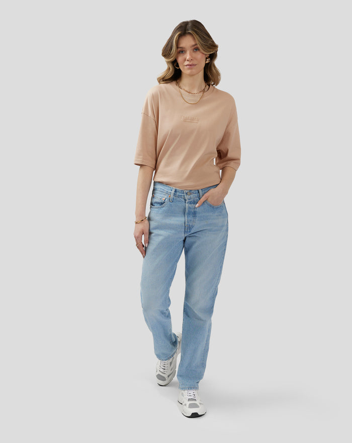 Woman in beige oversized short sleeve t shirt with embossed chest logo and back neck logo, paired with denim jeans