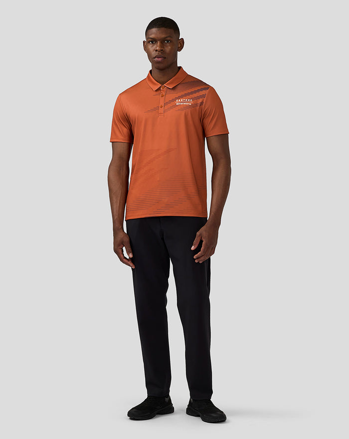 Full-body view of model in orange rust Castore Silverstone polo paired with black trousers and sneakers