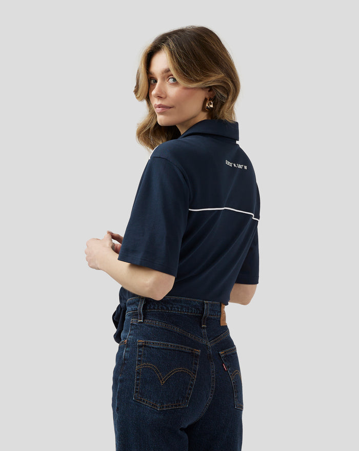 Three-quarter rear view of navy polo showing coordinates on the back and white piping along the back seam.