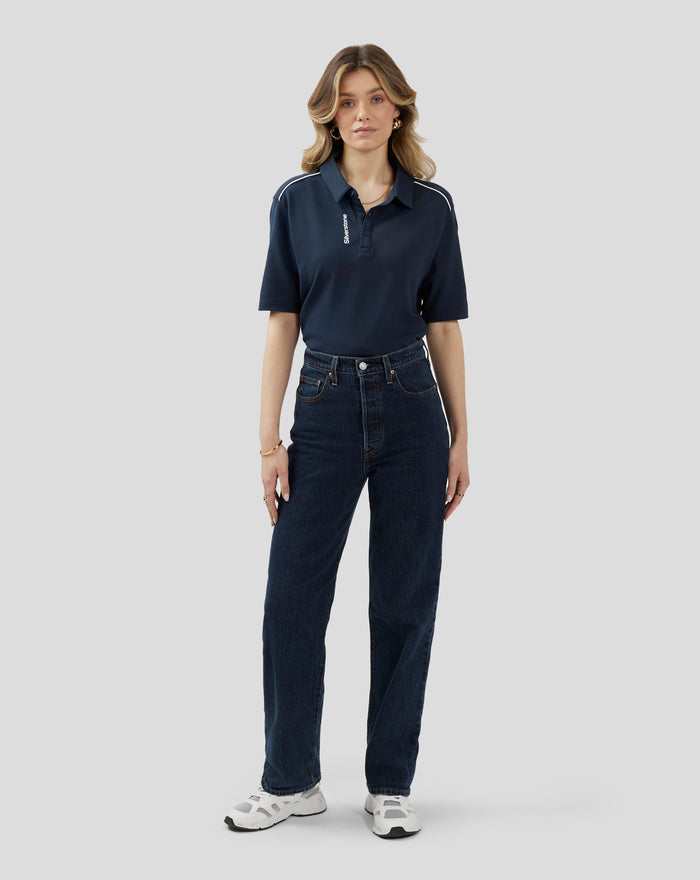 Full body shot of a woman in a navy polo with white piping and Silverstone branding on the placket, paired with jeans and white sneakers.
