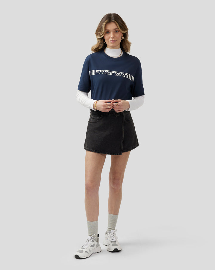Model wearing navy Silverstone Core T Shirt with white chest graphic, white underlayer, and black denim skirt.