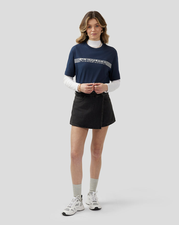 Model wearing navy Silverstone Core T Shirt with white chest graphic, white underlayer, and black denim skirt.