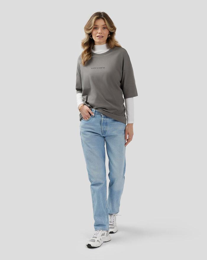 Model wearing charcoal grey oversized unisex T shirt with white underlayer, light blue jeans and sneakers; front silicone chest graphic.