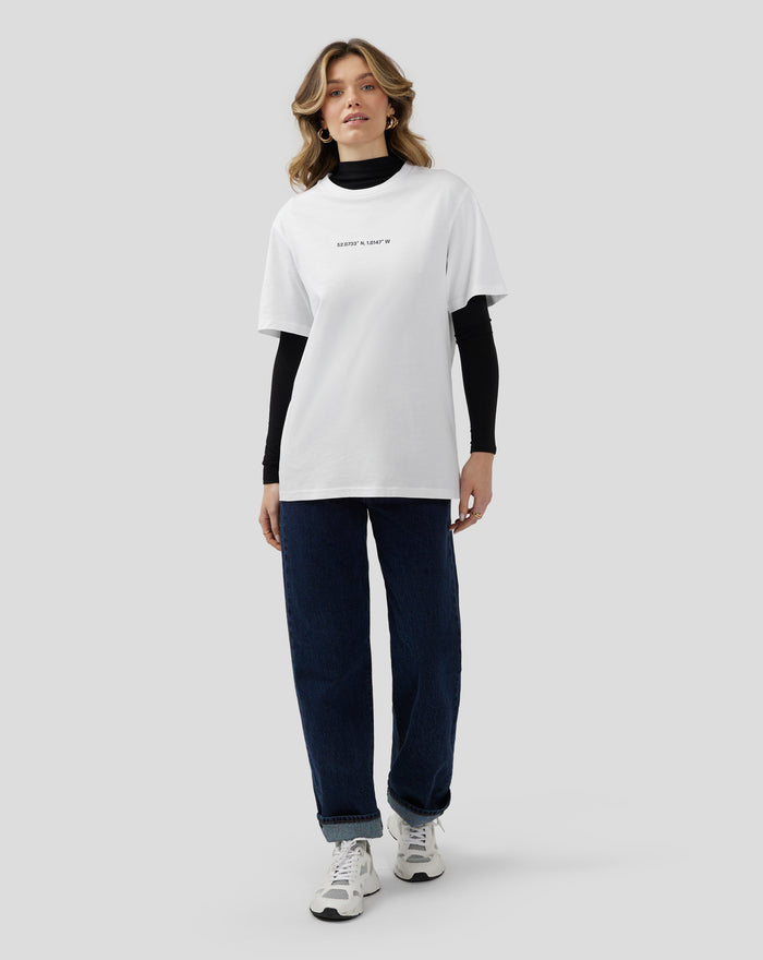 Full body view of a model in a white Silverstone Core Graphic T Shirt with coordinates printed on the chest paired with dark jeans and sneakers